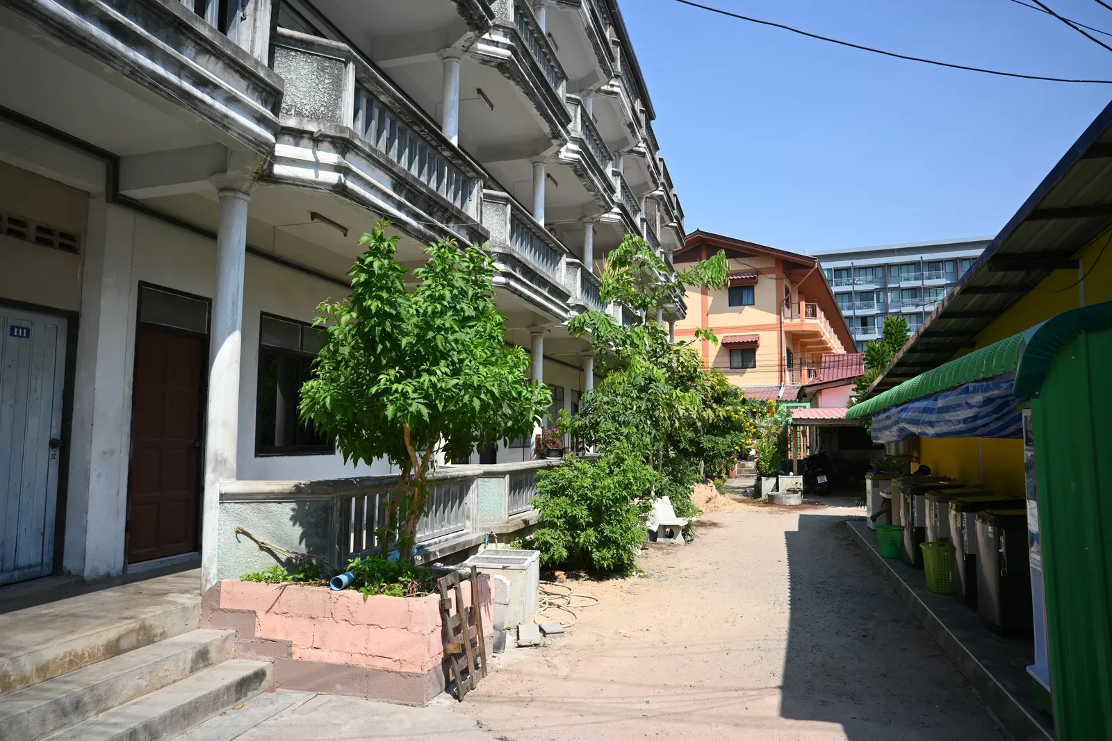 #5 - Sheltered Concrete in Tropical Urban Buildings (DFC_4541: Quiet sunlit alley between rows of low-rise buildings, lined with potted trees and shaded walkways., CC BY-SA 4.0)